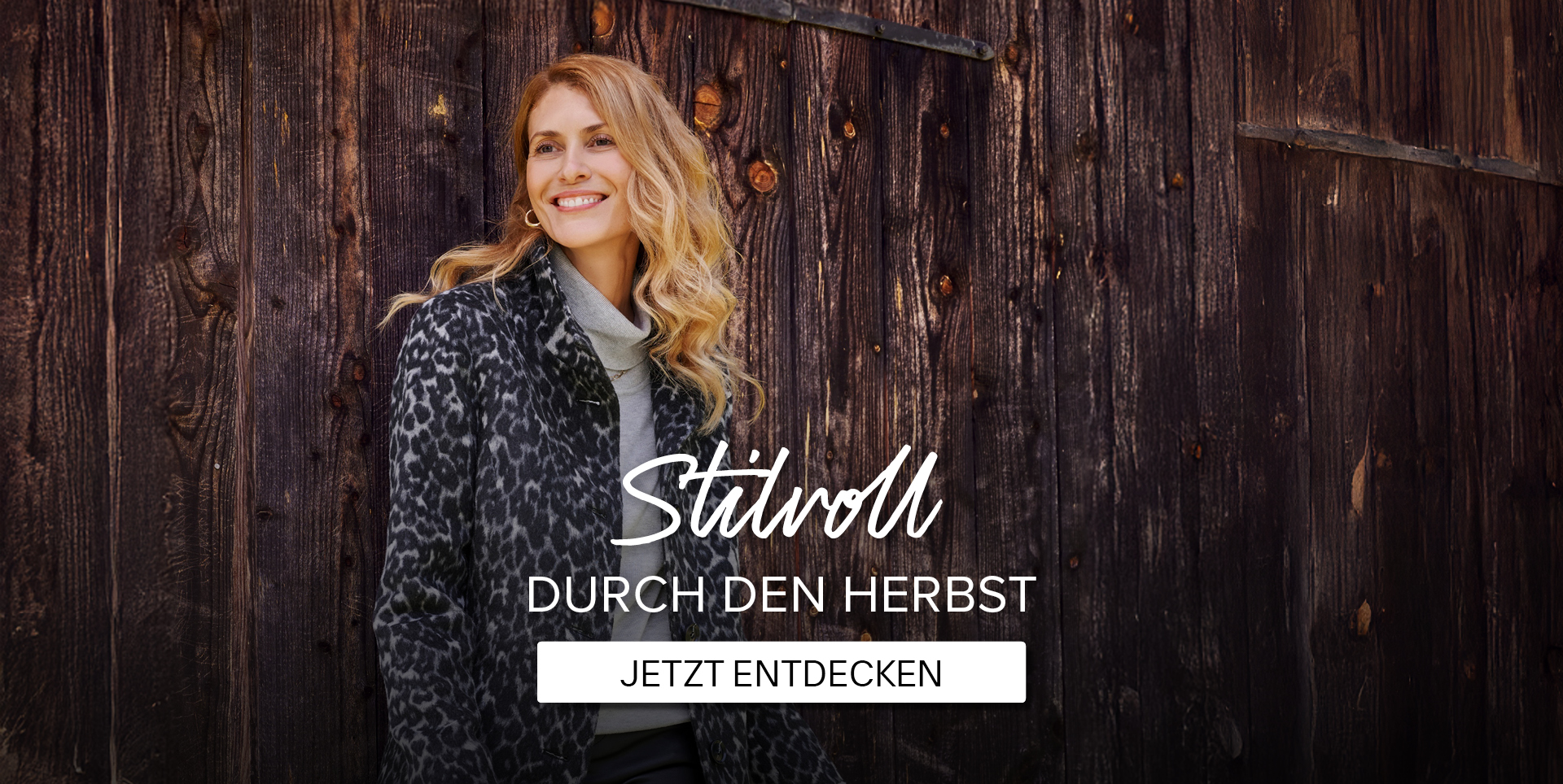 Eine Frau in einem modischen, leopardgemusterten Mantel steht an einer Holzwand. Text: "Stilvoll durch den Herbst - Jetzt entdecken."