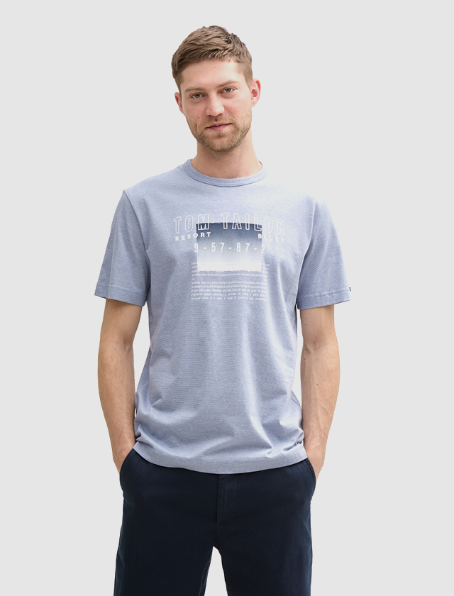 Ein Mann trägt ein hellblaues T-Shirt mit dem Schriftzug „Tom Tailor“ und sitzt vor einem neutralen Hintergrund. Ein Mann trägt ein hellblaues T-Shirt mit dem Schriftzug "Tom Tailor" und sitzt vor einem neutralen Hintergrund.