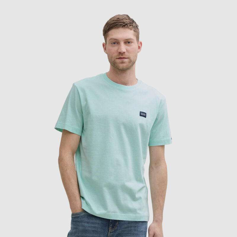 Ein Mann trägt ein mintgrünes T-Shirt und Jeans, steht vor einem neutralen Hintergrund.