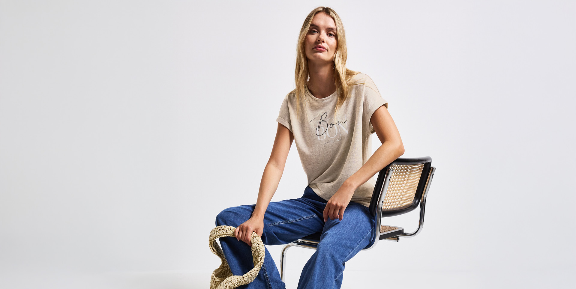 Eine Frau sitzt auf einem Stuhl, trägt ein hellbeiges T-Shirt mit dem Schriftzug „Bon“ und hat eine Tasche in der Hand. Eine Frau sitzt auf einem Stuhl, trägt ein hellbeiges T-Shirt mit dem Schriftzug "Bon" und hat eine Tasche in der Hand.