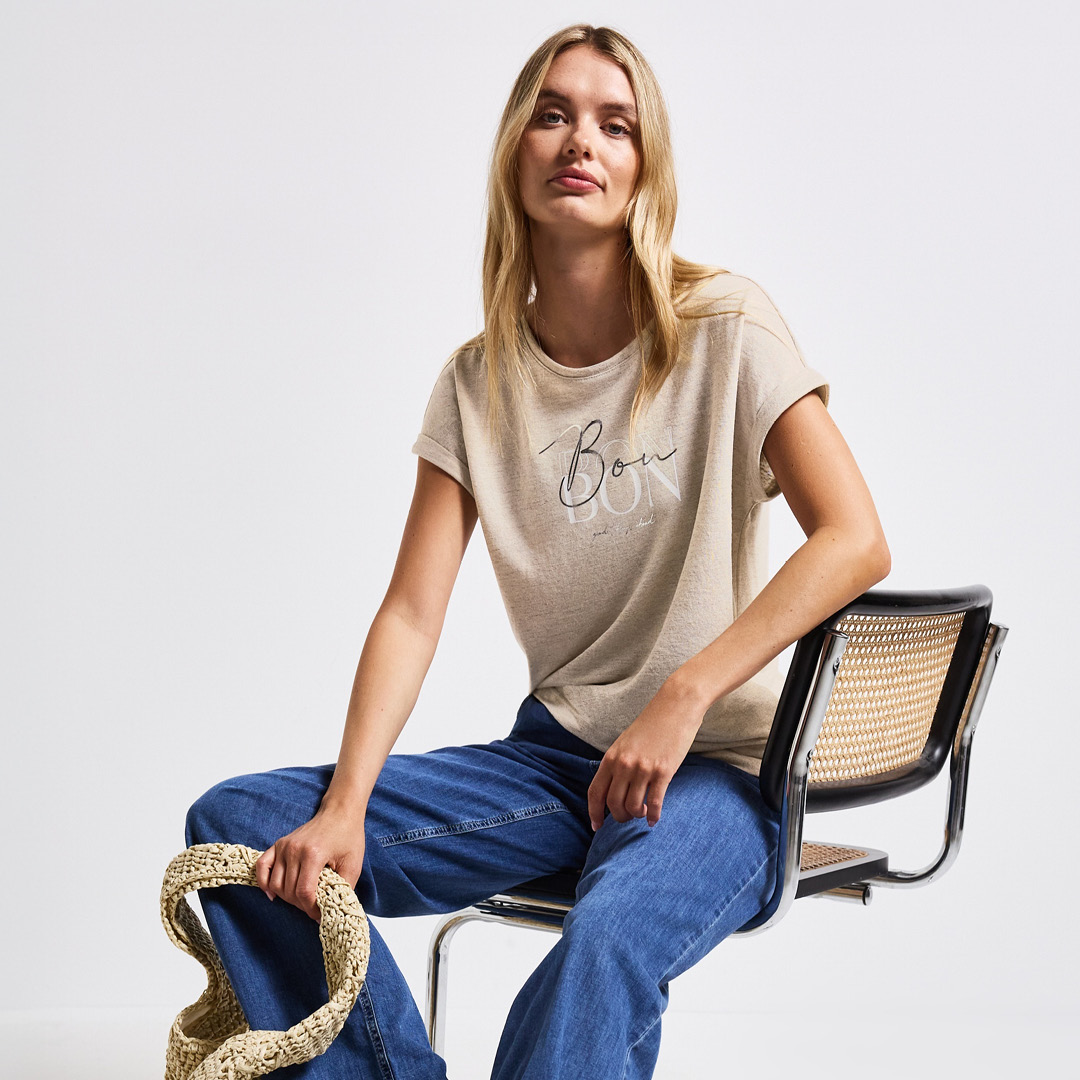 Eine Frau sitzt auf einem Stuhl, trägt ein hellbeiges T-Shirt mit dem Schriftzug „Bon“ und hat eine Tasche in der Hand. Eine Frau sitzt auf einem Stuhl, trägt ein hellbeiges T-Shirt mit dem Schriftzug "Bon" und hat eine Tasche in der Hand.