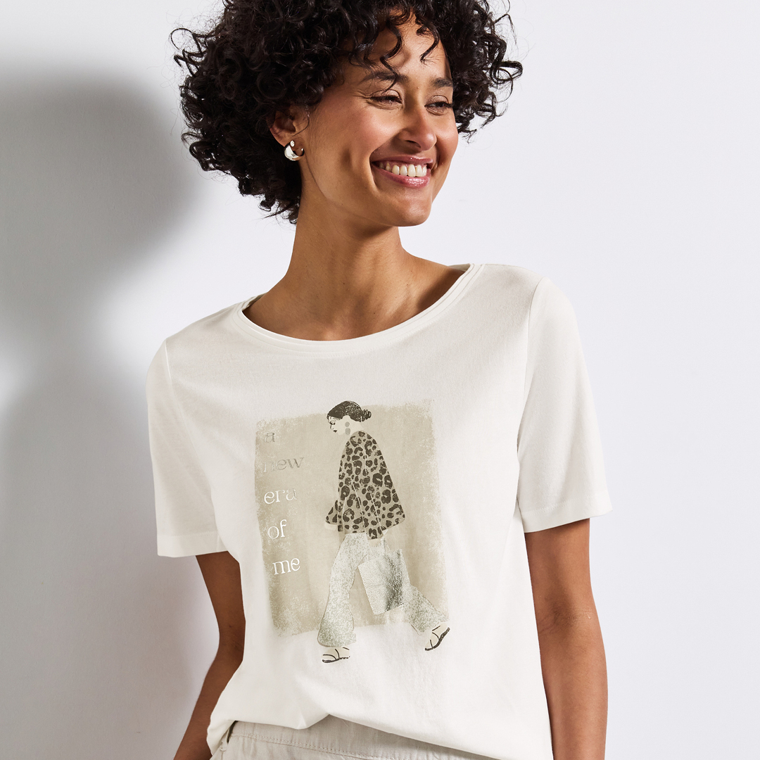 Eine lächelnde Frau mit lockigen Haaren trägt ein helles T-Shirt mit einem gezeichneten Motiv und einem Schriftzug.
