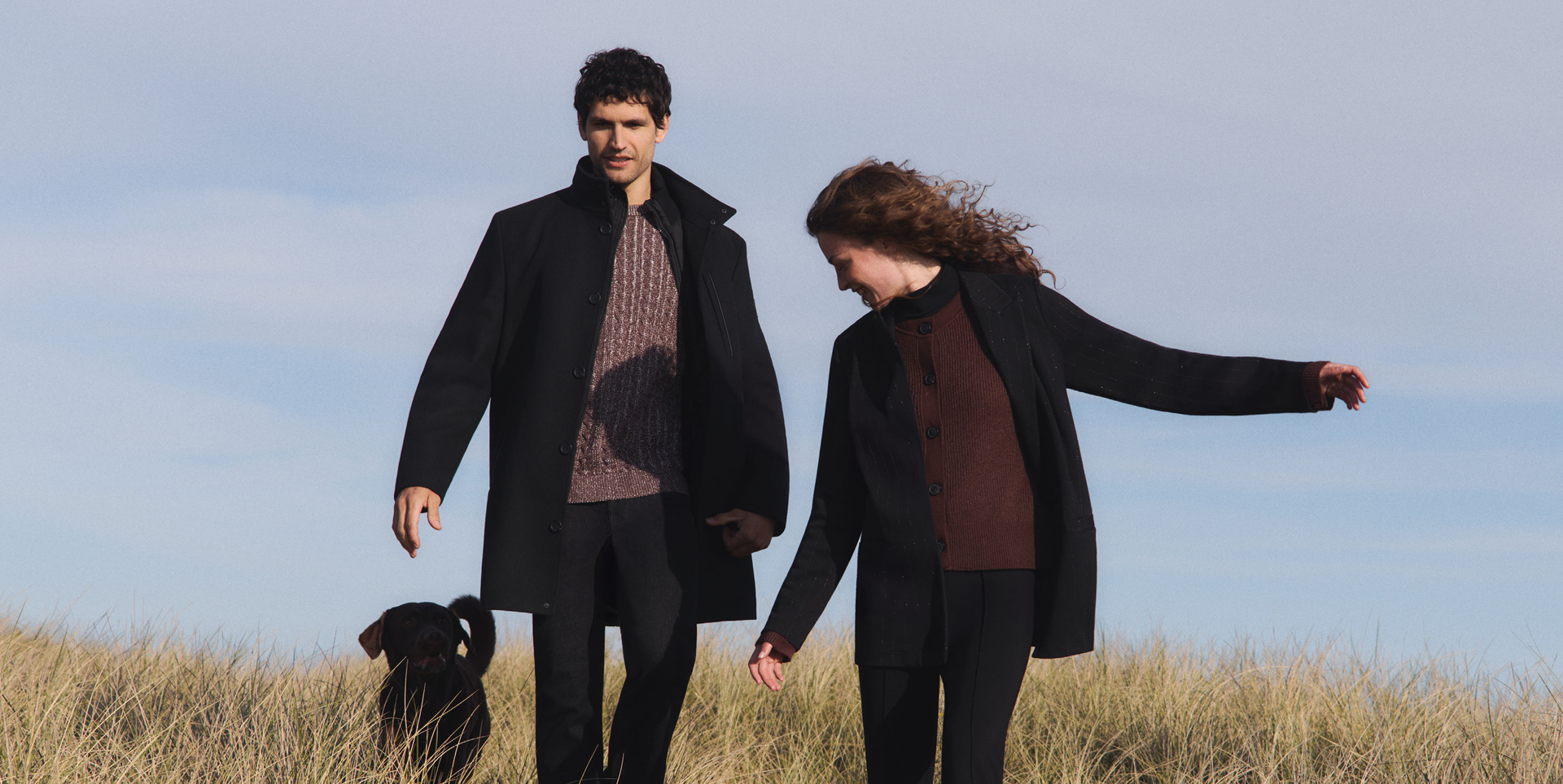 Zwei Personen in dunkler Oberbekleidung gehen durch hohes Gras, eine Person hält die Hand der anderen, ein schwarzer Hund läuft neben ihnen. Zwei Personen in dunkler Oberbekleidung gehen durch hohes Gras, eine Person hält die Hand der anderen, ein schwarzer Hund läuft neben ihnen.