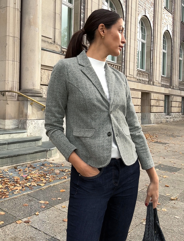 Frau trägt graues, tailliertes Sakko mit zwei Knöpfen, weiße Bluse und dunkle Jeans, steht vor historischer Fassade. Frau trägt graues, tailliertes Sakko mit zwei Knöpfen, weiße Bluse und dunkle Jeans, steht vor historischer Fassade.