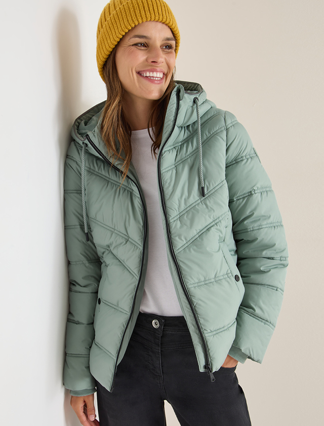 Person trägt eine hellgrüne gesteppte Jacke mit Kapuze, weiße T-Shirt und schwarze Hose, gelbe Strickmütze Person trägt eine hellgrüne gesteppte Jacke mit Kapuze, weiße T-Shirt und schwarze Hose, gelbe Strickmütze