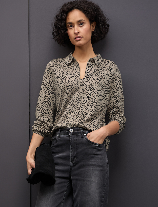 Person trägt eine beige Bluse mit schwarzem Tupfenmuster und schwarze Jeans, hält schwarzen Stoff in der linken Hand. Person trägt eine beige Bluse mit schwarzem Tupfenmuster und schwarze Jeans, hält schwarzen Stoff in der linken Hand.